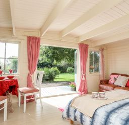 A room with a brown leather sofa a bed with cups on a tray a table with two chairs and red curtains at Songbird Hideaway in Pwllheli