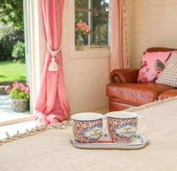 Two cups on a tray on a table with a blanket near a leather sofa and a window with red curtains at Songbird Hideaway in Pwllheli
