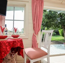 A dining area with a red tablecloth on a round table set with plates and wine glasses next to a white chair with a red checkered cushion at Songbird Hideaway Pwllheli