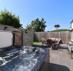 An outdoor patio with a hot tub a dining table with chairs and a cushioned sofa at Rosebank Cottage in Mortehoe