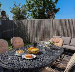 An outdoor patio with a metal dining table and cushioned chairs with fruit and cake on the table and a sofa against a wooden fence at Rosebank Cottage in Mortehoe