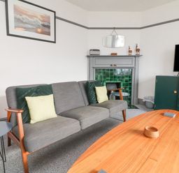 A living room with a gray sofa green cushions a wooden coffee table a fireplace with green tiles a dresser and a television at Chilton in Bude