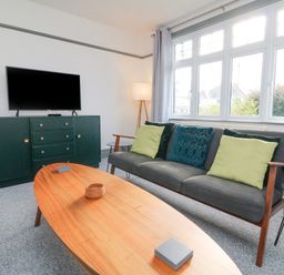 A living room with a gray sofa with green and blue cushions a wooden coffee table a dark green cabinet with a television and a floor lamp at Chilton in Bude