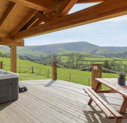 A wooden deck with a hot tub and picnic table overlooking green hills at Bryn Eiddon Log Cabin in Machynlleth