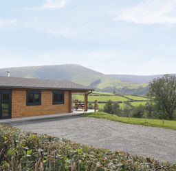 A wooden log cabin with a gravel driveway and a picnic bench on a porch with hills and trees in the background at Bryn Eiddon Log Cabin in Machynlleth