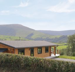 A wooden cabin with a gravel path and hedge in front with hills in the background at Bryn Eiddon Log Cabin in Machynlleth