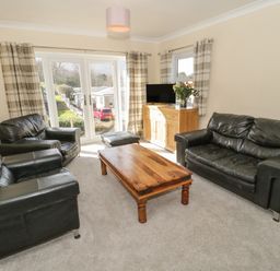 A living room with black leather sofas a wooden coffee table a wooden cabinet with a television flowers and checked curtains at Arwelfa in Penmaenmawr