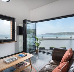 A sitting area with a leather sofa and orange cushions next to a wooden coffee table and a balcony overlooking the beach at The Old Beach Store in Sennen Cove