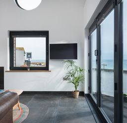 A living room with a fireplace a wall mounted TV a plant a window and glass doors showing a sea view at The Old Beach Store in Sennen Cove