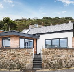 A modern house with stone walls wooden cladding and large windows by a hillside at The Old Beach Store in Sennen Cove