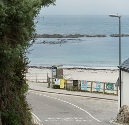 A coastal road with signs by a wooden fence and a sandy beach with ocean in the background at The Old Beach Store in Sennen Cove