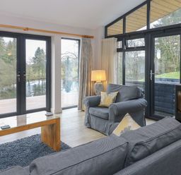 A living room with grey sofas a wooden coffee table a lamp and a fireplace with glass doors overlooking trees and a river at Tarn Lodge in Bowness-On-Windermere