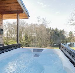 An outdoor hot tub on a wooden deck overlooking trees and a small pond at Tarn Lodge in Bowness-On-Windermere