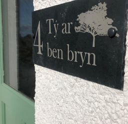 A house number plaque with the text 4 Ty ar ben bryn and a tree symbol on a textured white wall next to a green door at Ty ar Ben Bryn in Pencarnisiog near Rhosneigr