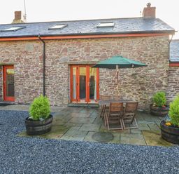 A stone exterior patio with wooden table and chairs under a green umbrella surrounded by planter barrels at Little Brook Cottage in Laugharne