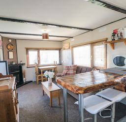 A living room with a wooden table and chairs a brown sofa with pillows a wooden coffee table with flowers a wooden sideboard a microwave and kitchen counter with stools at Castle View Cabin in Cloughton