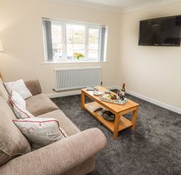 A living room with a beige sofa, wooden coffee table, wall-mounted TV, and window with a flower vase at Station View in Whitby