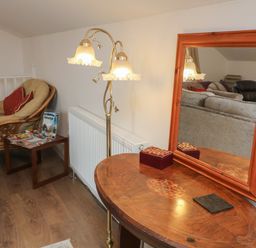 A corner with a cushioned wicker chair next to a small table with magazines a wooden table with a framed mirror and a floor lamp at Scenic Views in High Bickington