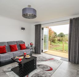 A living room with a gray sofa with red pillows a black coffee table with red mugs and pastries gray curtains open to a garden at Rosamond in Shipton Gorge