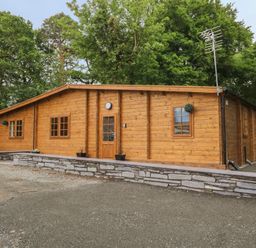 An exterior view of a wooden cabin surrounded by trees with a paved area and a stone retaining wall at Bryn Derwen Lodge in Bethesda