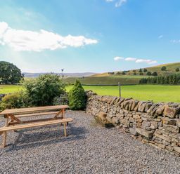 A wooden picnic table on a gravel area next to a stone wall with green fields and hills in the background at Field Barn in Alston