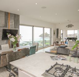 An open living space with a dining table, kitchen island with gas stove, chairs, flowers on tables, a television on a stone wall, large windows with a view of the sea, a hanging chair, and a foosball table at Hafod Y Mor in Criccieth