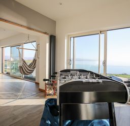 A room with a foosball table and a hanging hammock chair with large windows showing a sea view at Hafod Y Mor in Criccieth