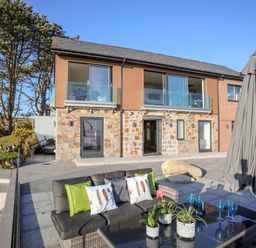 An outdoor patio with a sofa, table with plants and glasses, umbrella, and a two-story house with stone and wood exterior at Hafod Y Mor in Criccieth