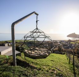 A metal hanging chair with cushions on a grassy hill overlooking a coastal town and sea at Hafod Y Mor in Criccieth