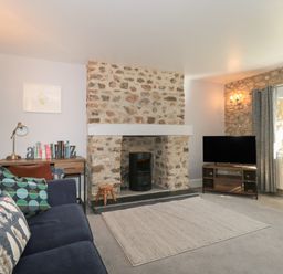 A living room with a sofa a desk with books a stone fireplace a TV on a stand and a window with curtains at The Linhay in Chardstock