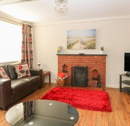 A living room with a brown leather sofa red and beige cushions a red rug brick fireplace and a TV on a glass stand at Compass Cottage in Bodham near Sheringham