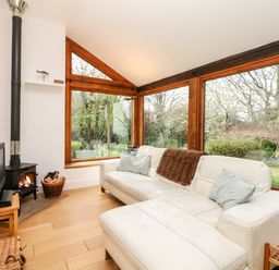 A living room with a white sofa, wood burning stove, television on a wooden stand, and large windows showing a garden at Puffin Cottage in Hawkchurch
