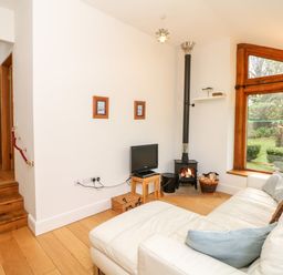A living room with a white sofa and pillows a small television on a wooden stand a wood-burning stove and a large window showing a garden at Puffin Cottage in Hawkchurch