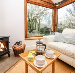 A corner of a living room with a wood burning stove a basket with firewood a small wooden table with tea items and a sofa with a striped cushion at Puffin Cottage in Hawkchurch