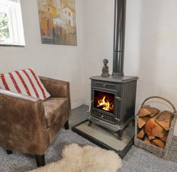A leather armchair with a cushion beside a wood burning stove with fire and a basket of firewood at Little House in Red Wharf Bay