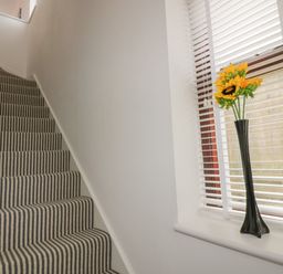 A staircase with striped carpet and a window with blinds and a vase of sunflowers on the windowsill at Elin Howlyek in Newquay