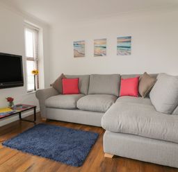 A living room with a grey sectional sofa with red and brown cushions a blue rug a wall mounted TV and small windows at Elin Howlyek in Newquay