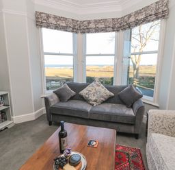 A living room with a grey sofa and a wooden coffee table in front of bay windows showing a rural view at Whinstones at 4 The Villas in Embleton