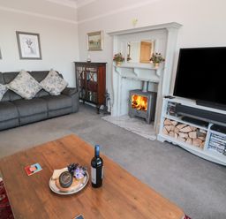 A living room with a grey sofa and floral cushions a wooden coffee table a fireplace with a wood stove and a TV on a white stand filled with wood and DVDs at Whinstones at 4 The Villas in Embleton
