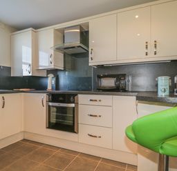 A kitchen with white cabinets a built-in oven a microwave coffee accessories and a green bar stool at Rowan Cottage in St Bees