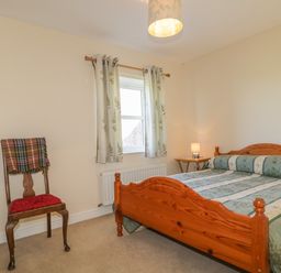 A bedroom with a wooden bed and green patterned bedspread a wooden chair with a plaid blanket and window with patterned curtains at Rowan Cottage in St Bees