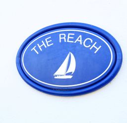 A blue oval sign with a white sailboat and the words The Reach on a white wall at The Reach in Hallsands
