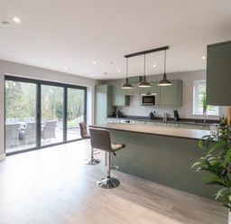 A kitchen with green cabinets an island with two bar stools pendant lights and sliding glass doors leading to an outdoor seating area at Fron Haul - Robin Goch Llanfwrog near Ruthin