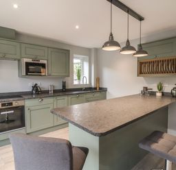 A kitchen with green cabinets a countertop with two chairs and hanging lights at Fron Haul - Robin Goch in Llanfwrog near Ruthin