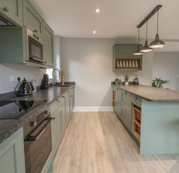 A kitchen with green cabinets a gray countertop a stovetop an oven a microwave hanging lights and bar stools at Fron Haul - Robin Goch in Llanfwrog near Ruthin