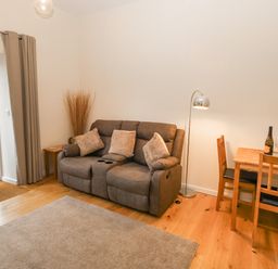 A living room with a brown sofa and three cushions a wooden table with two chairs a floor lamp and a vase with flowers at Tyddyn Sianel - Derw in Llanystumdwy near Criccieth