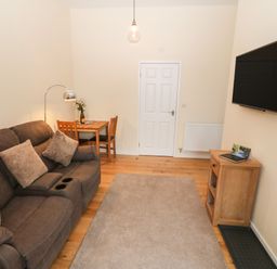 A living room with a brown sofa, wooden dining table with chairs, wall-mounted TV, and wooden cabinet at Tyddyn Sianel - Derw in Llanystumdwy near Criccieth