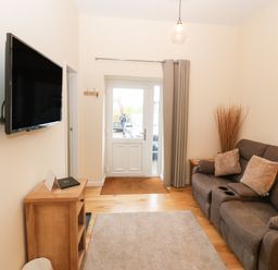 A living room with a gray sofa, wall-mounted TV, wooden cabinet, rug, and a door with a curtain at Tyddyn Sianel - Derw in Llanystumdwy near Criccieth