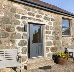 Stone exterior wall with a dark gray door two wooden benches and flower pots at Tyddyn Sianel - Derw in Llanystumdwy near Criccieth