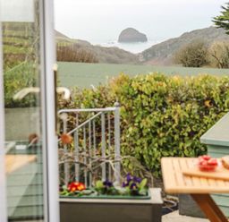 A view of a garden with a hedge and hills beyond with a rock in the sea seen through an open door with a wooden table holding bread and a glass of wine at Glenhaven in Trewarmett near Tintagel